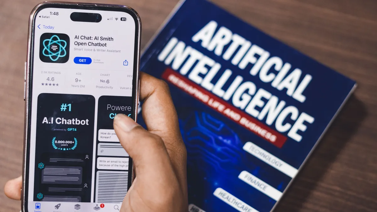 A hand holding a cell phone with an AI chatbot app open, alongside a book titled "Artificial Intelligence" on a wooden surface.