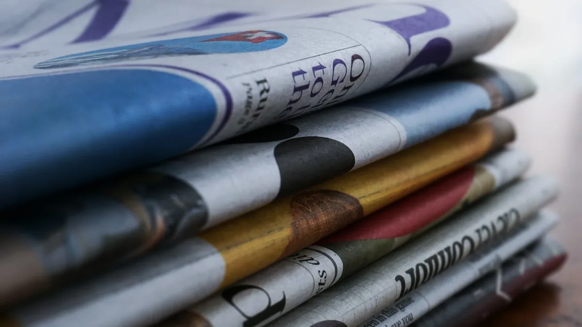 A stack of newspapers with various headlines and advertisements visible.